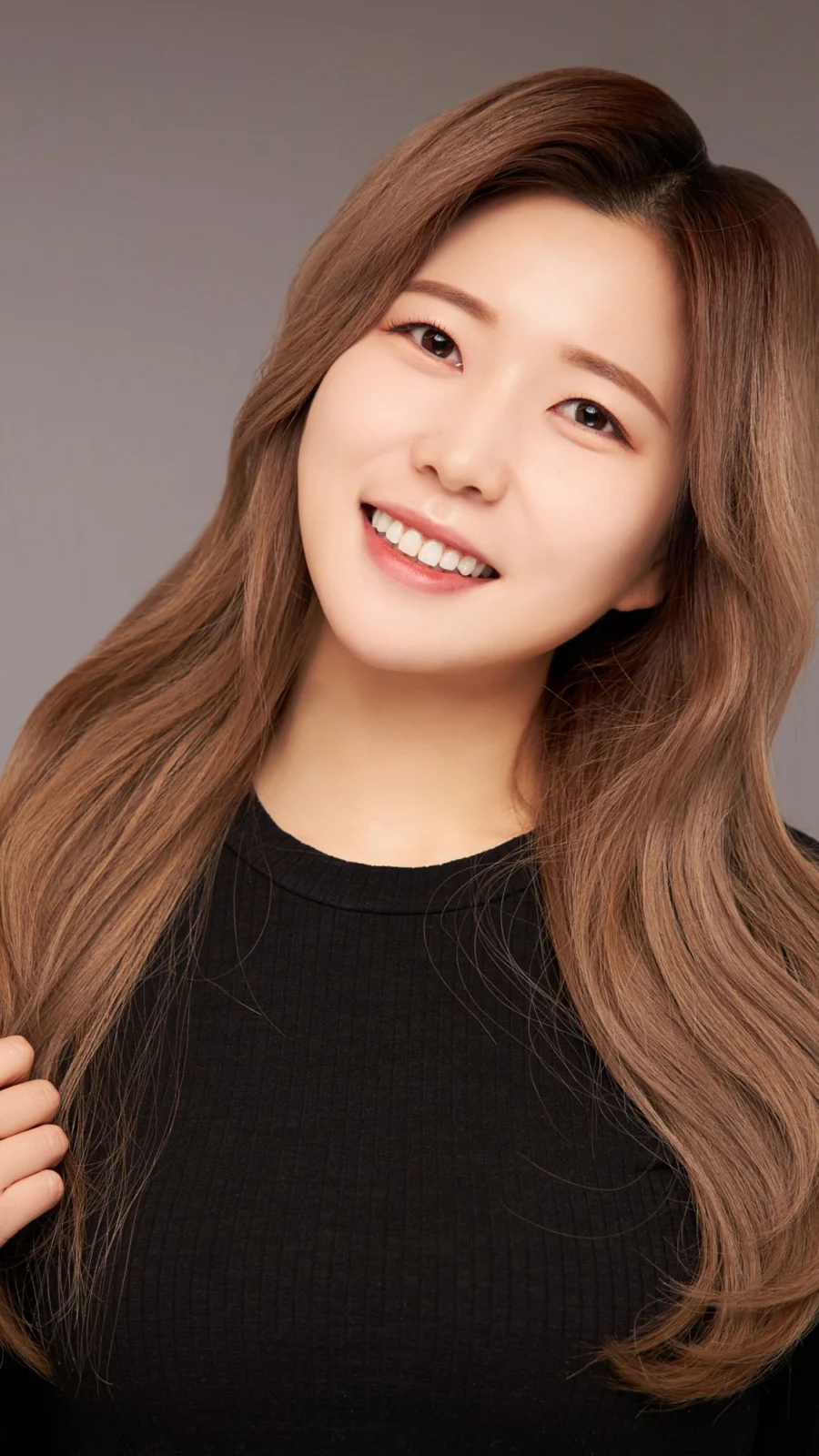 회색 배경에서 갈색 긴 머리를 넘기며 웃는 검정 니트 차림 프로필 사진