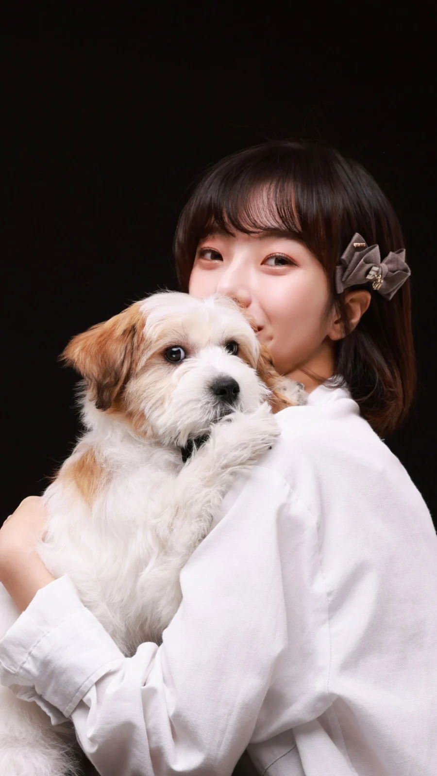 검정 배경에서 흰색과 갈색 털 강아지를 안고 볼을 댄 한 사람의 개족사진