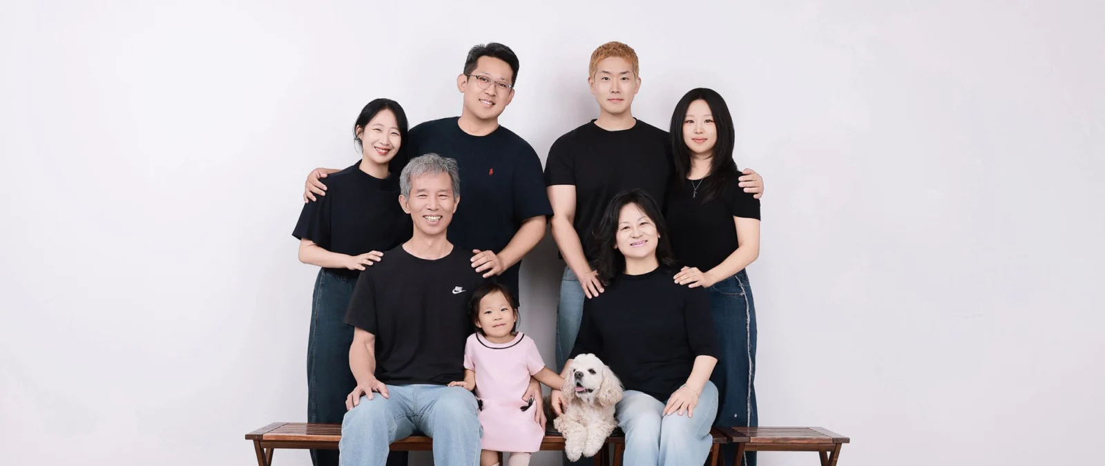 벤치에 앉은 세 명 뒤로 네 명이 선 검은 티셔츠 가족과 흰 강아지 세로 개족사진
