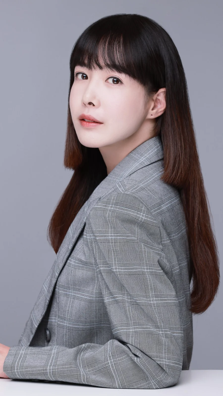 회색 배경에서 체크 재킷 차림으로 테이블에 기대 앉은 프로필 사진