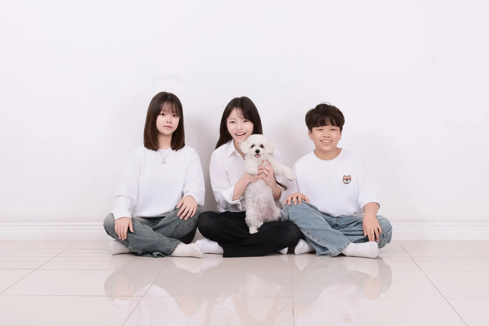 흰 바닥에 책상다리로 앉아 흰 강아지를 안은 세 명의 개족사진