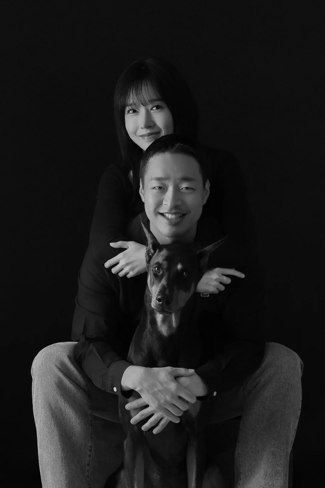 어두운 배경에서 검은 대형견을 안고 겹쳐 앉은 두 사람의 흑백 개족사진