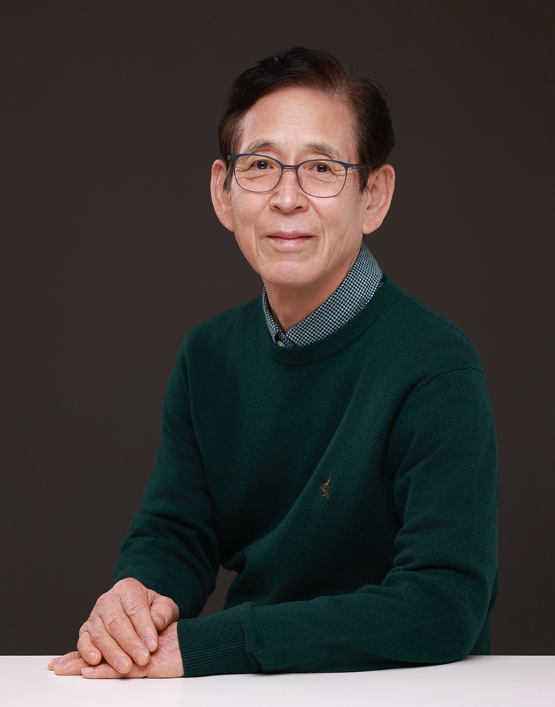 안경을 쓰고 초록 니트를 입은 인물이 흰 테이블에 손을 얹은 독사진