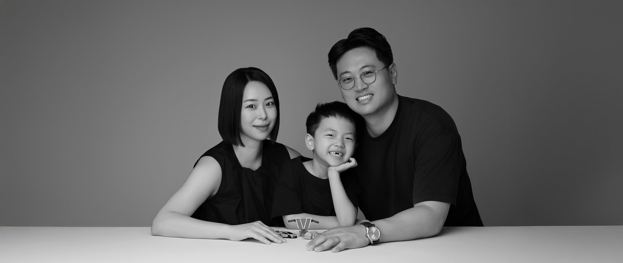 회색 배경 테이블 앞에서 턱을 괸 아이와 어른 두 명의 흑백 가족사진