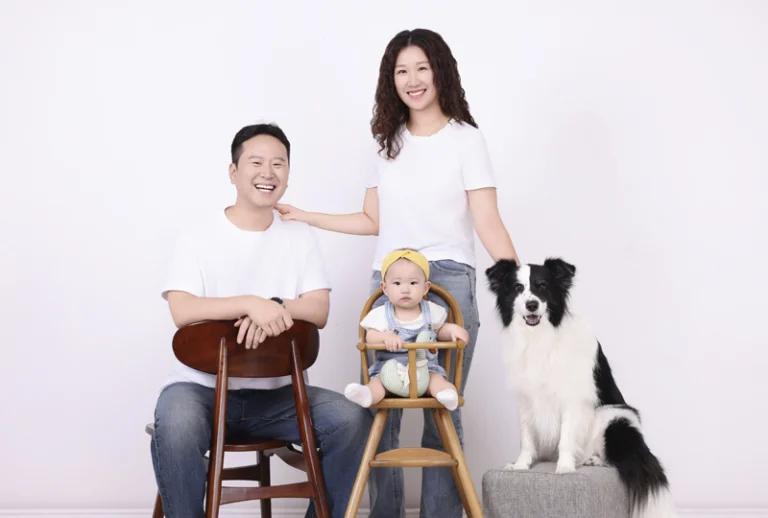 흰 배경 앞 흰 티셔츠 차림의 세 사람과 스툴 위에 앉은 흑백 강아지