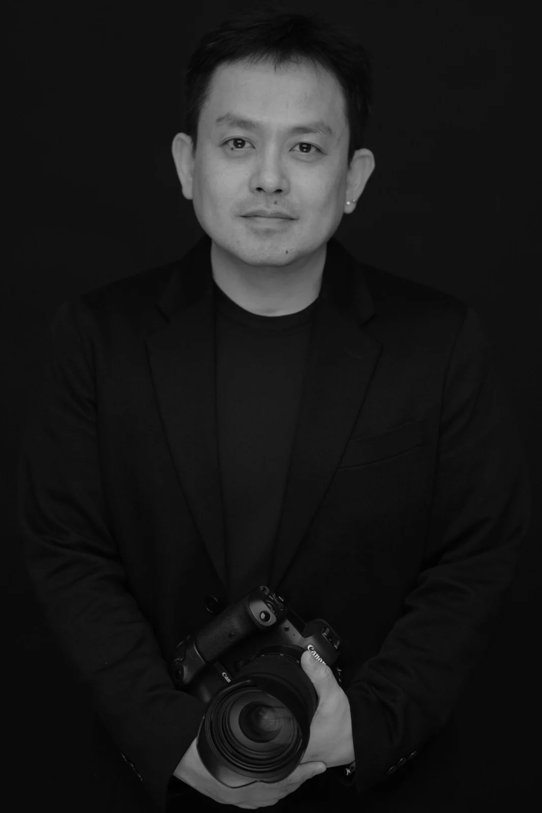 검은 배경 앞 검은 재킷 차림으로 카메라를 두 손에 든 인물의 흑백 사진