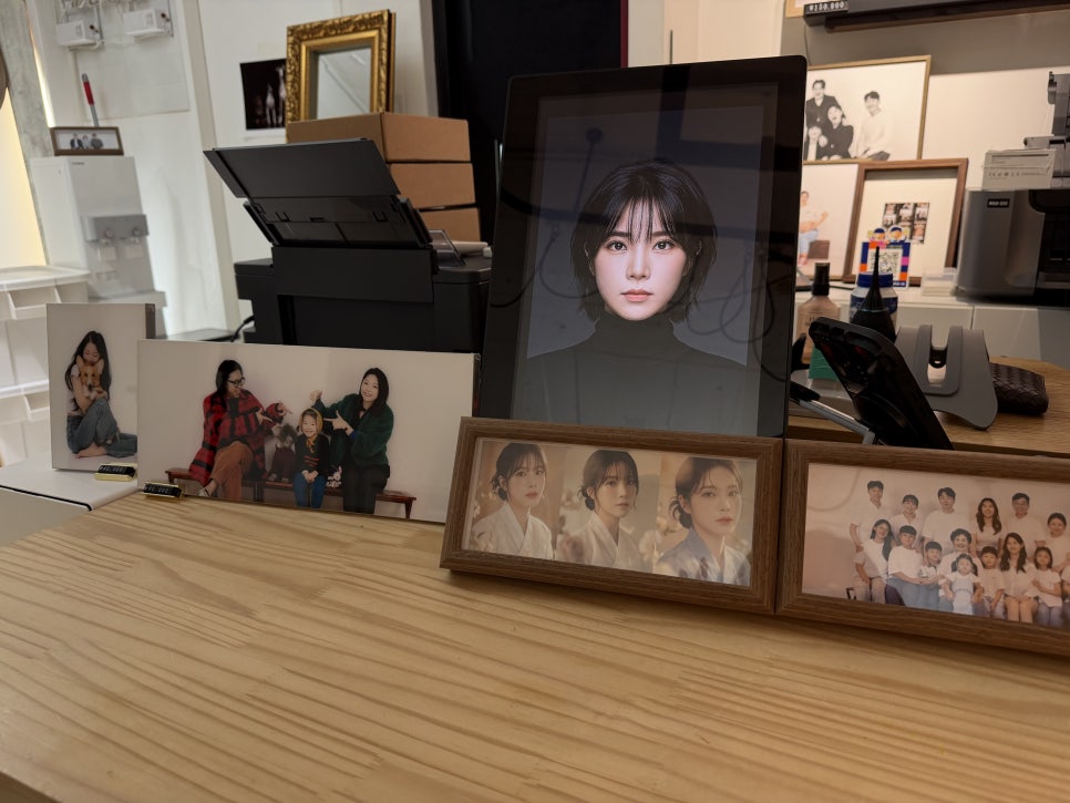 나무 책상 위 디지털 액자와 증명·가족 사진 액자들, 뒤로 프린터가 보이는 모습