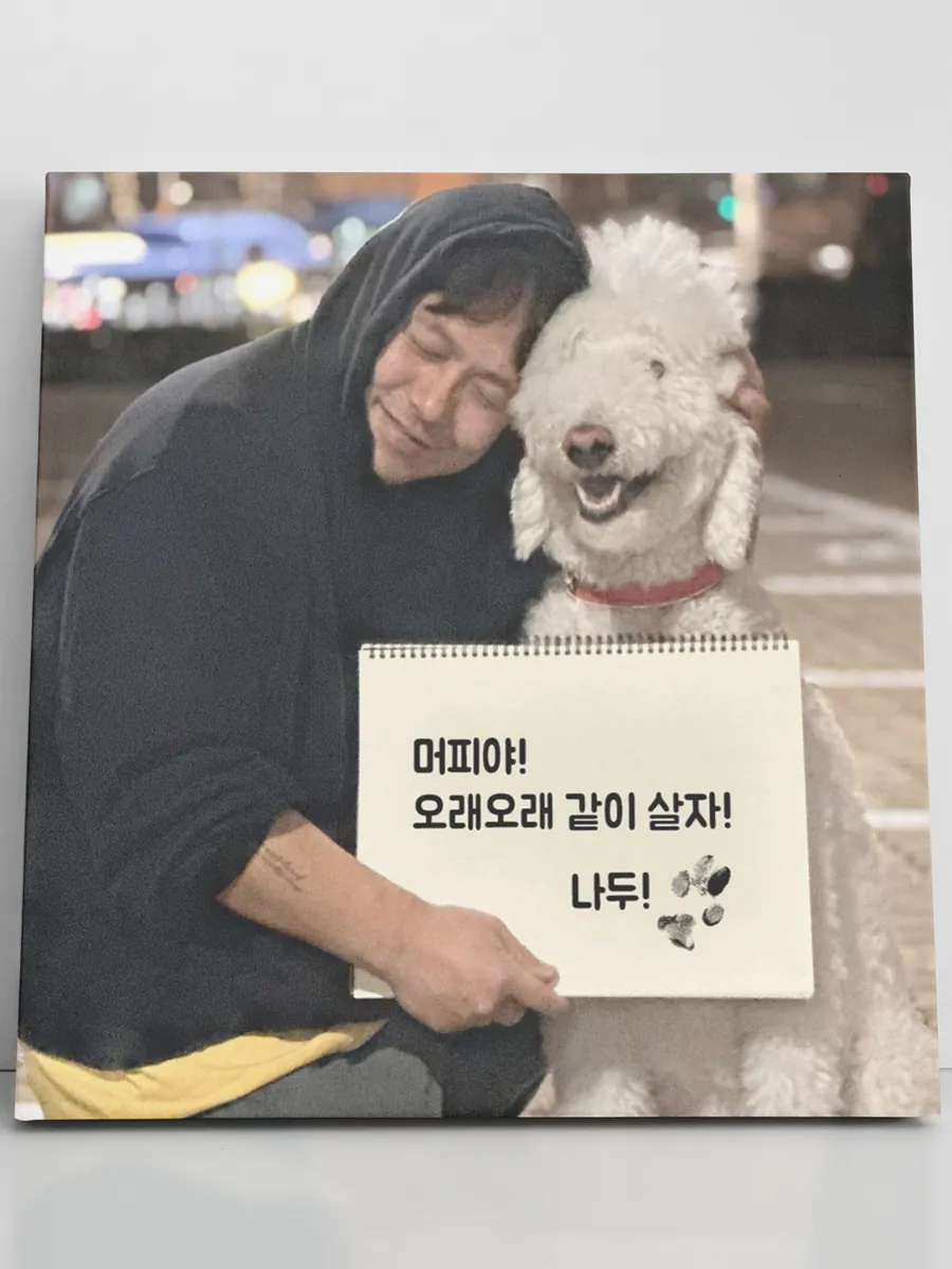검은 후드 차림의 인물이 흰 곱슬 강아지와 볼을 맞대고 발도장 메시지를 든 캔버스
