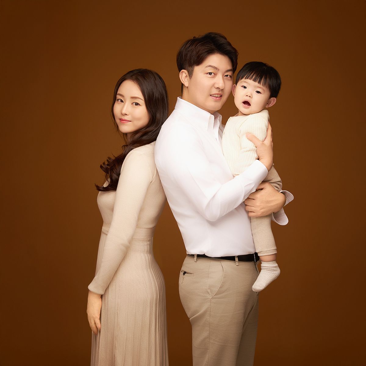 갈색 배경 앞 베이지와 흰색 옷을 맞춰 입은 세 사람, 한 명은 품에 안겨 있음