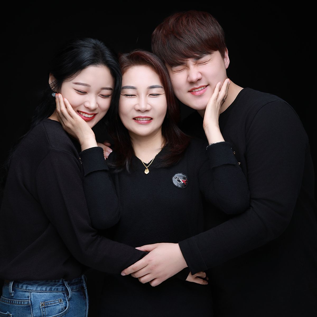 검은 배경 앞 검은 옷을 맞춰 입고 가운데 사람을 감싸 안은 세 사람