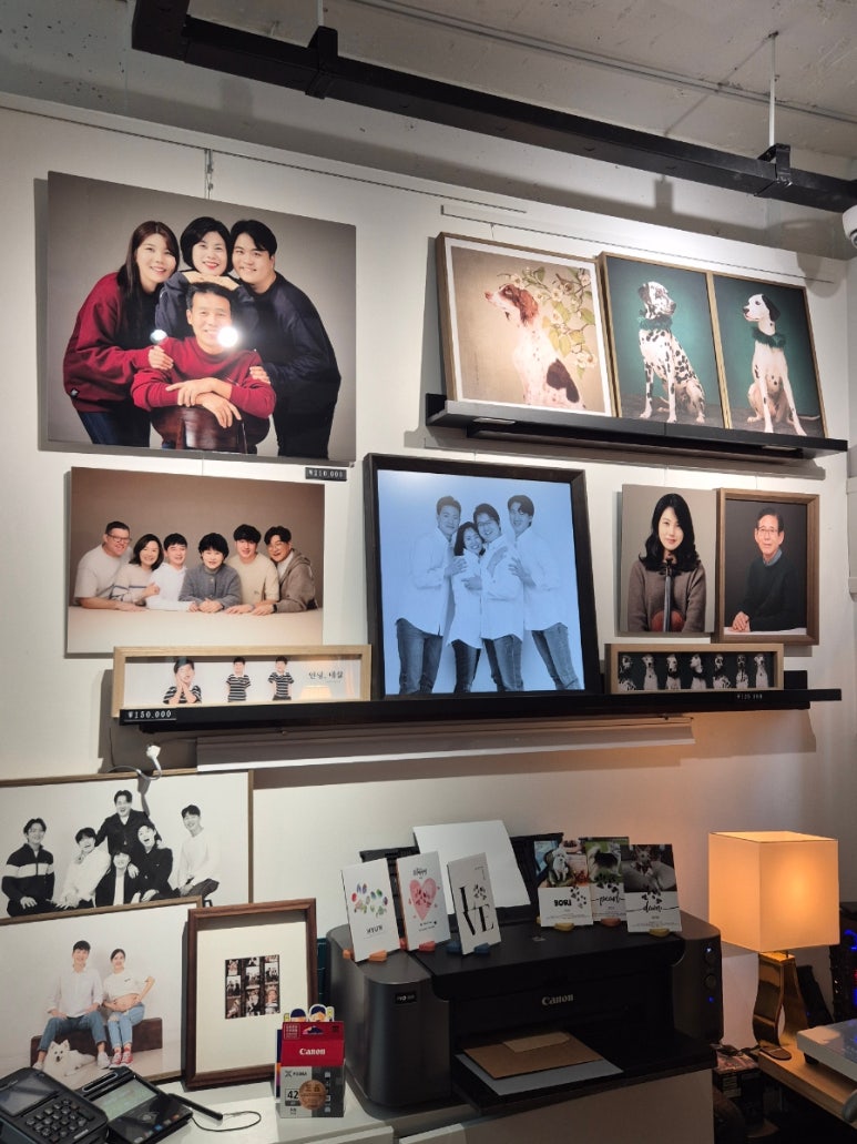 가족사진·반려견 액자가 벽 선반 가득 걸리고 아래 프린터가 놓인 스튜디오 벽면