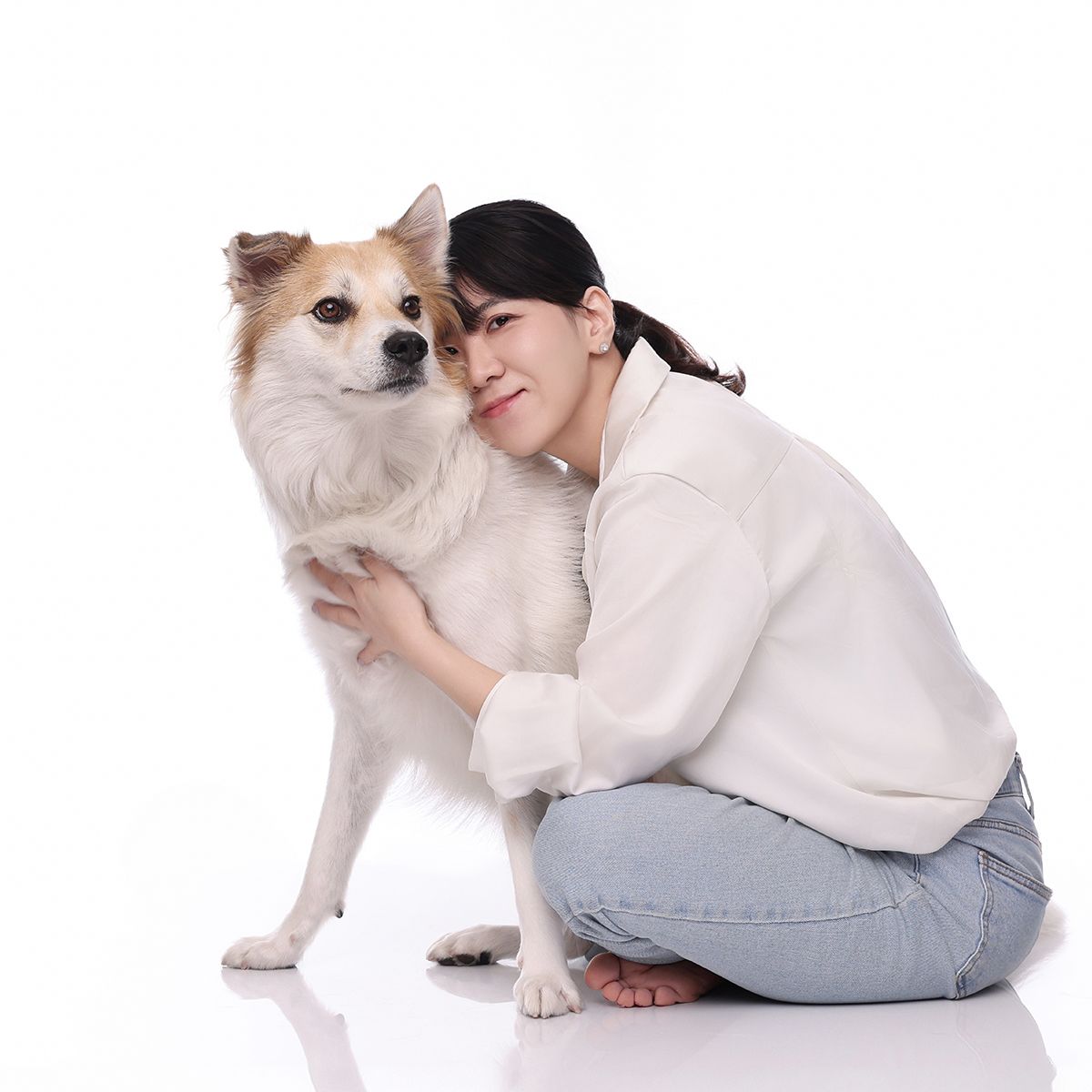 흰 셔츠와 청바지 차림으로 바닥에 앉아 흰 털에 갈색 귀 강아지를 안은 사진