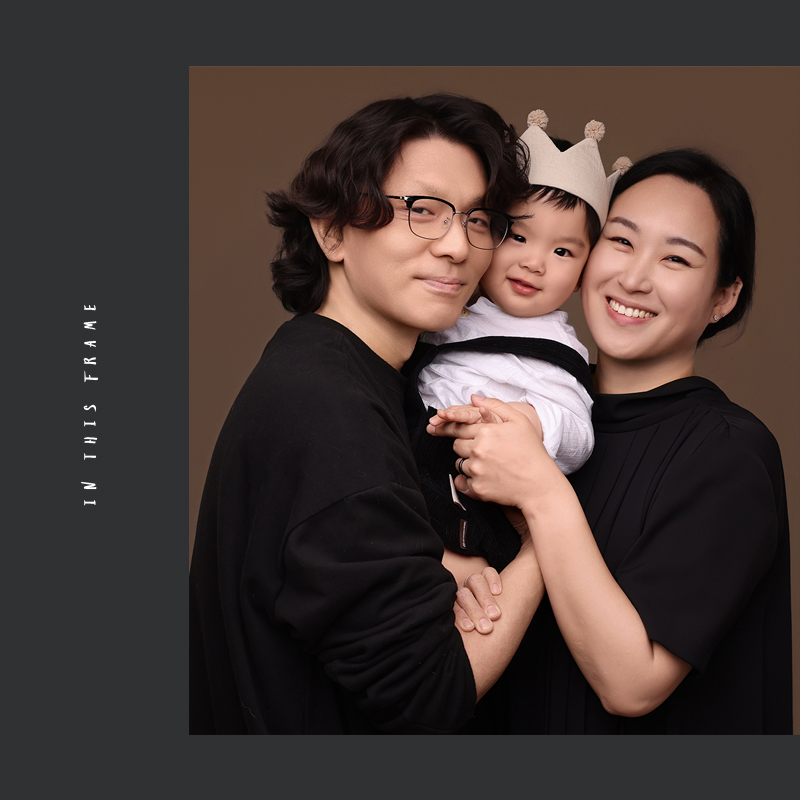 갈색 배경 앞 검은 옷의 두 사람이 왕관 모자를 쓴 한 명을 안은 사진, 영문 글자
