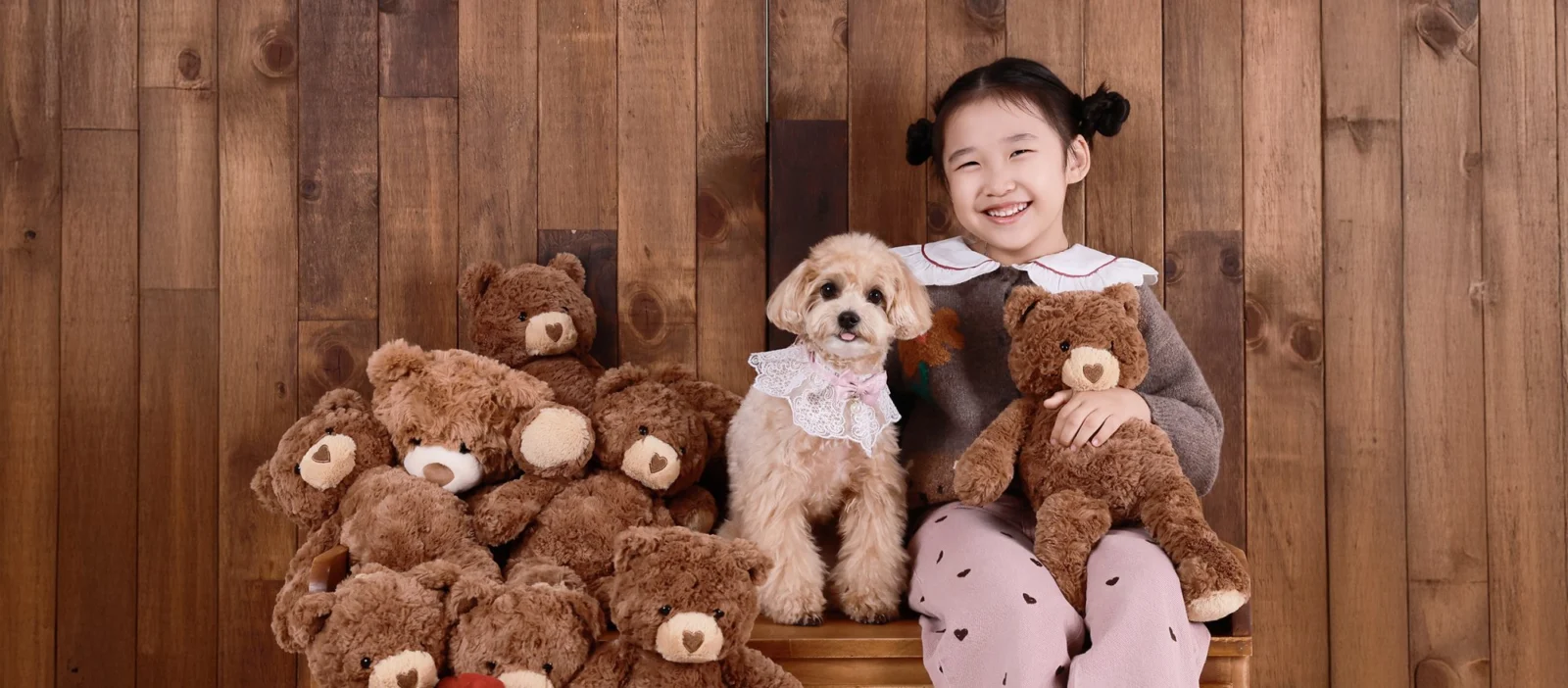 나무 벽 앞에서 곰 인형과 갈색 곱슬털 강아지 옆에 앉아 웃는 아이의 가로 개족사진