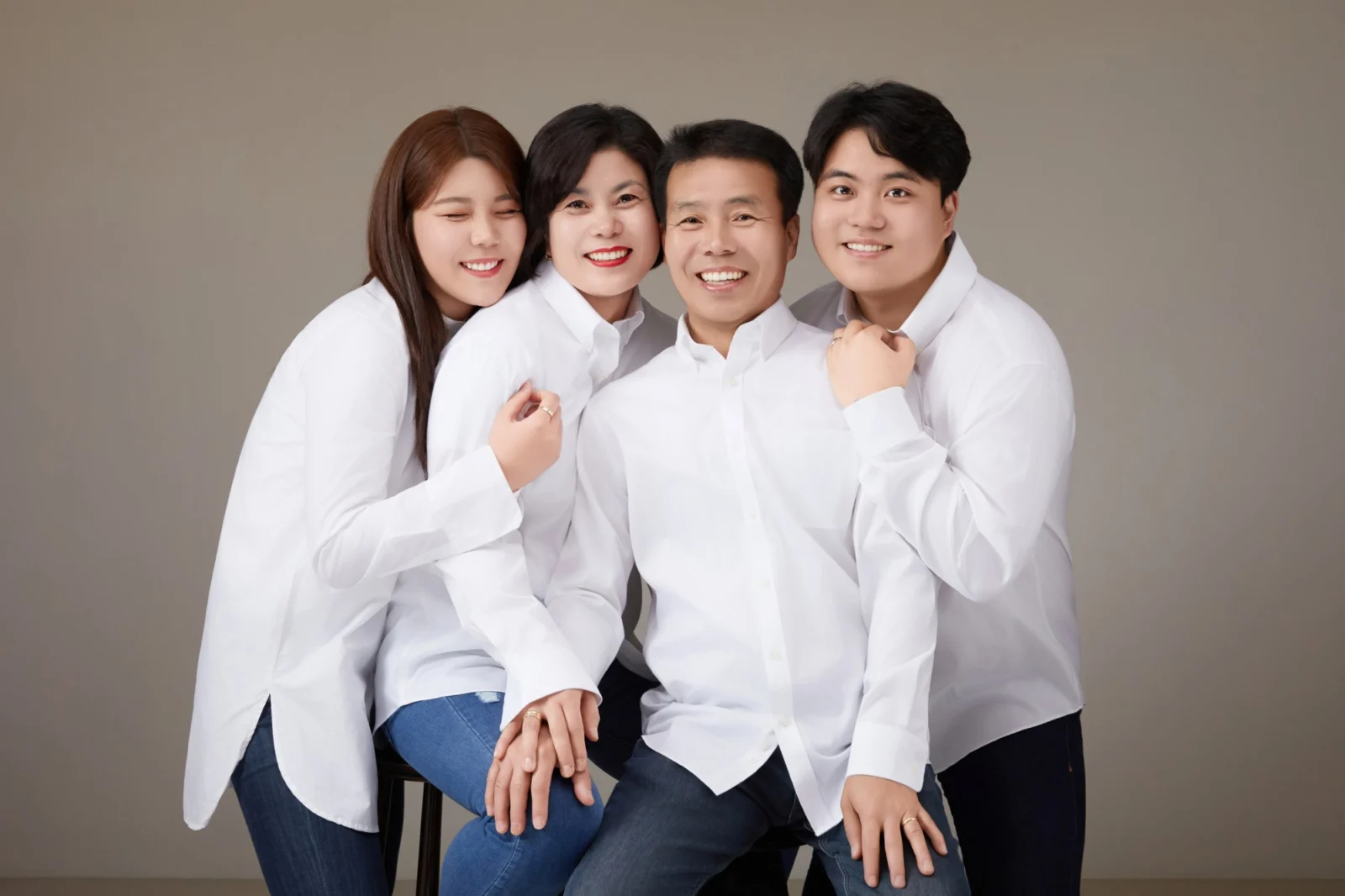 회색 배경에서 흰 셔츠를 맞춰 입고 기대어 앉은 네 명의 가족사진