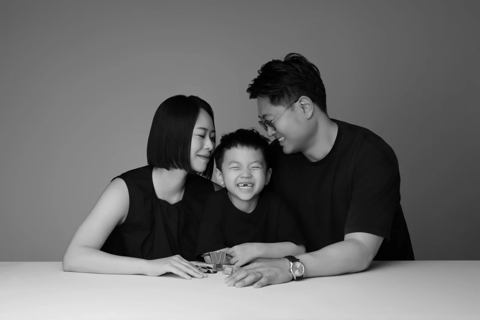 아이를 사이에 두고 얼굴을 기대어 웃는 어른 두 명의 흑백 가족사진