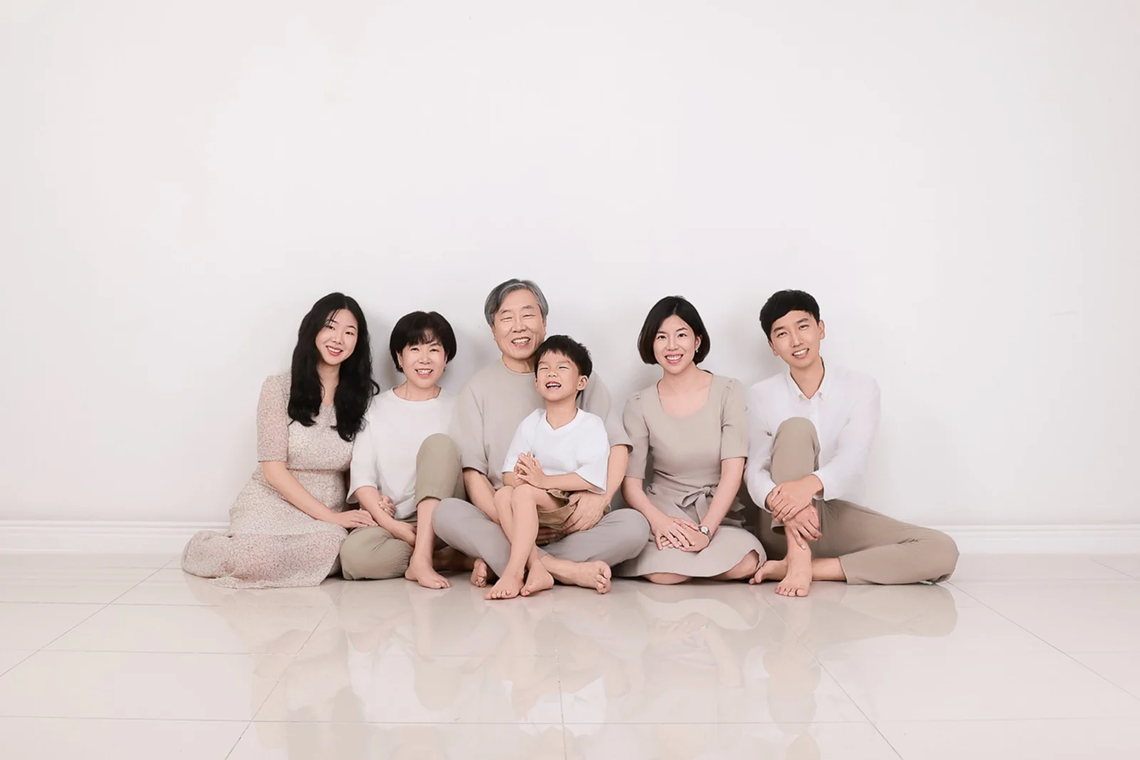 흰 배경 바닥에 베이지 톤 옷으로 앉은 여섯 명을 넓게 담은 가족사진