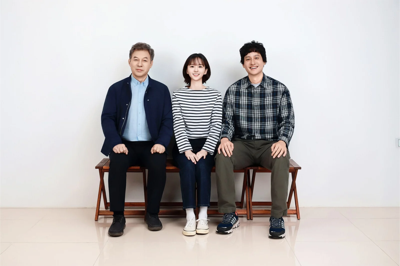 흰 배경 나무 벤치에 나란히 앉은 세 사람을 넓게 담은 가족사진