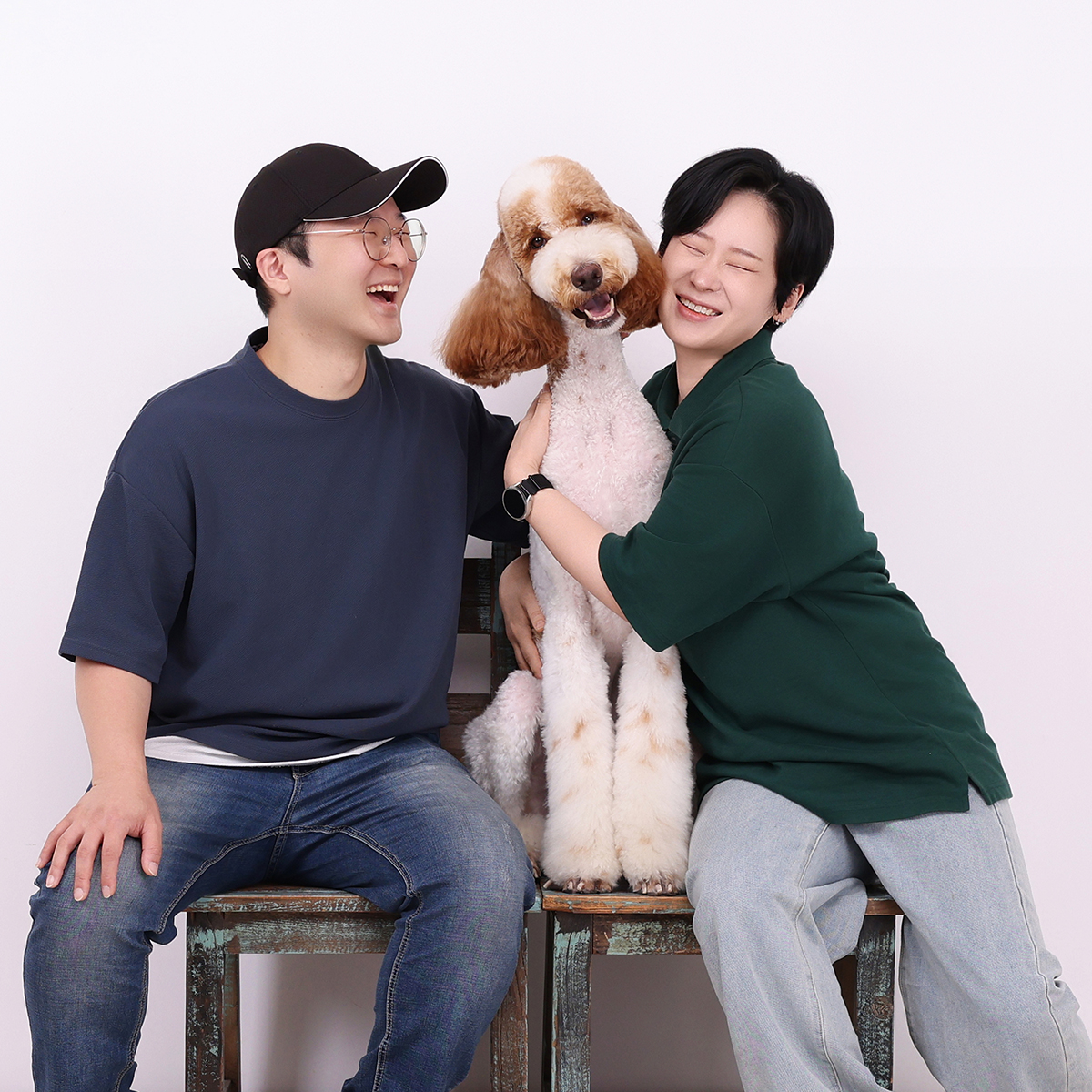 나무 의자에 앉은 두 사람이 사이에 앉은 큰 곱슬털 강아지를 안고 웃는 사진
