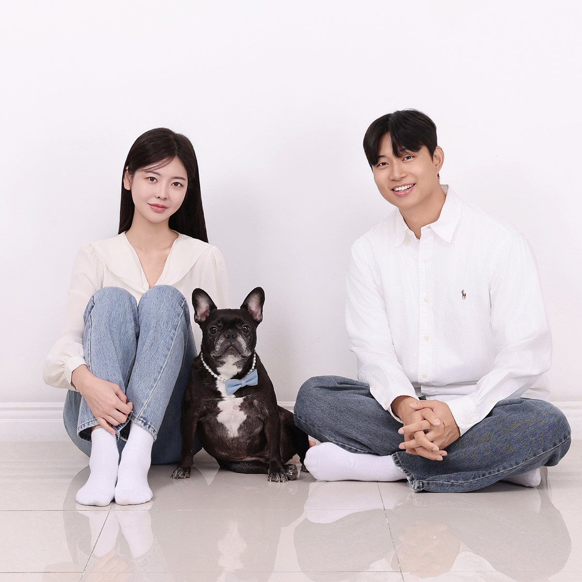 흰 벽 앞 바닥에 앉은 두 사람 사이로 나비넥타이를 한 검은 강아지