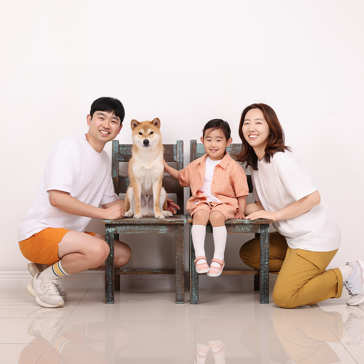 나무 의자 두 개에 앉은 황갈색 강아지와 한 명, 양옆에 무릎 꿇은 두 사람