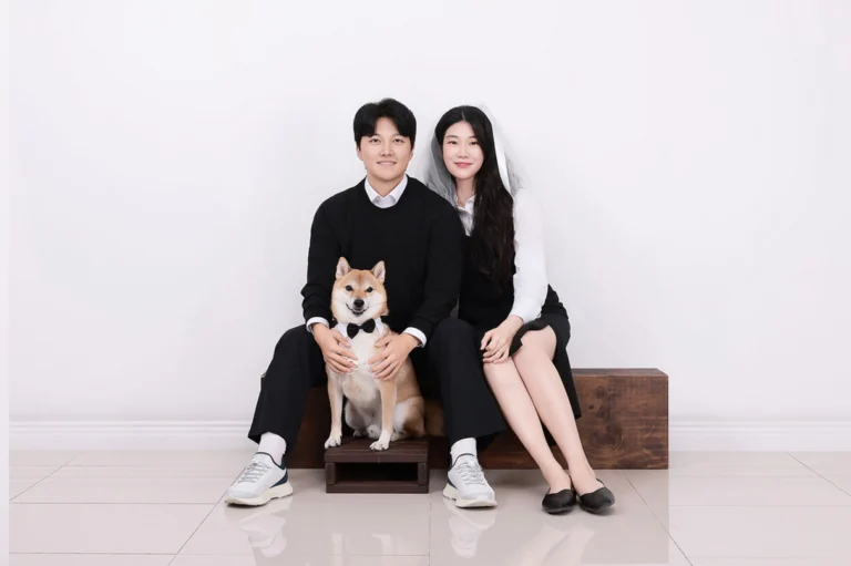 나무 단에 앉은 두 사람과 나비넥타이를 한 황갈색 강아지, 한 명은 면사포 차림
