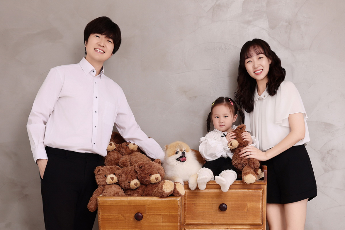 곰 인형 가득한 나무 서랍장 위에 앉은 한 명과 흰 강아지, 양옆에 선 두 사람