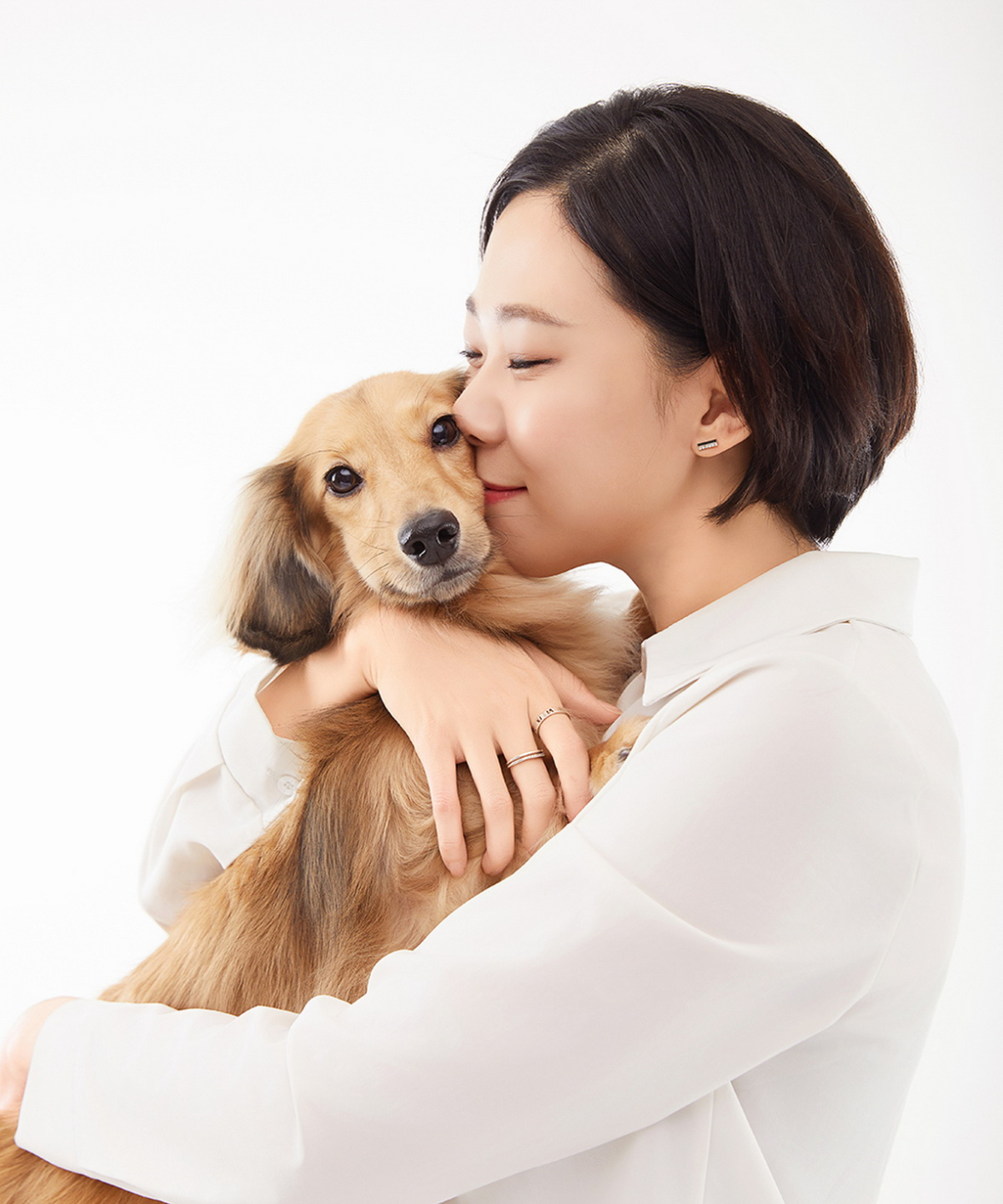 흰 셔츠 차림의 단발 인물이 눈을 감고 긴 털의 갈색 강아지에 볼을 댄 사진