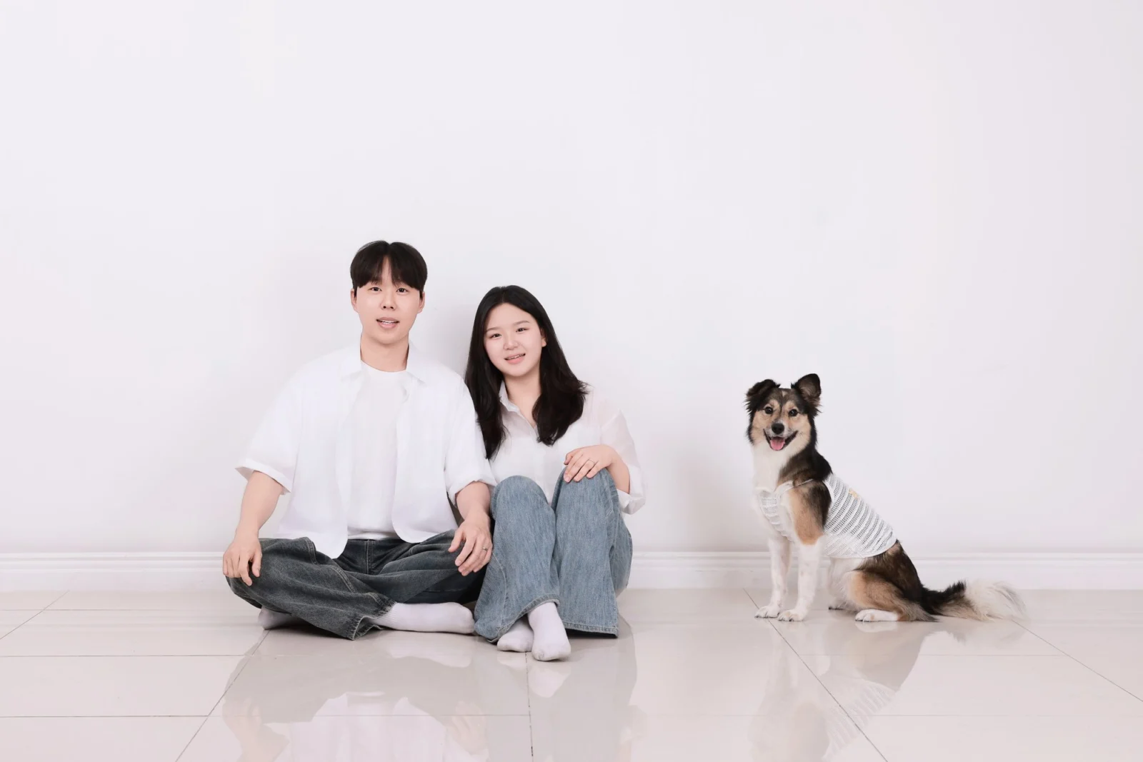 흰 바닥에 앉은 두 사람과 옆에 따로 앉은 세 가지 털색 강아지의 개족사진