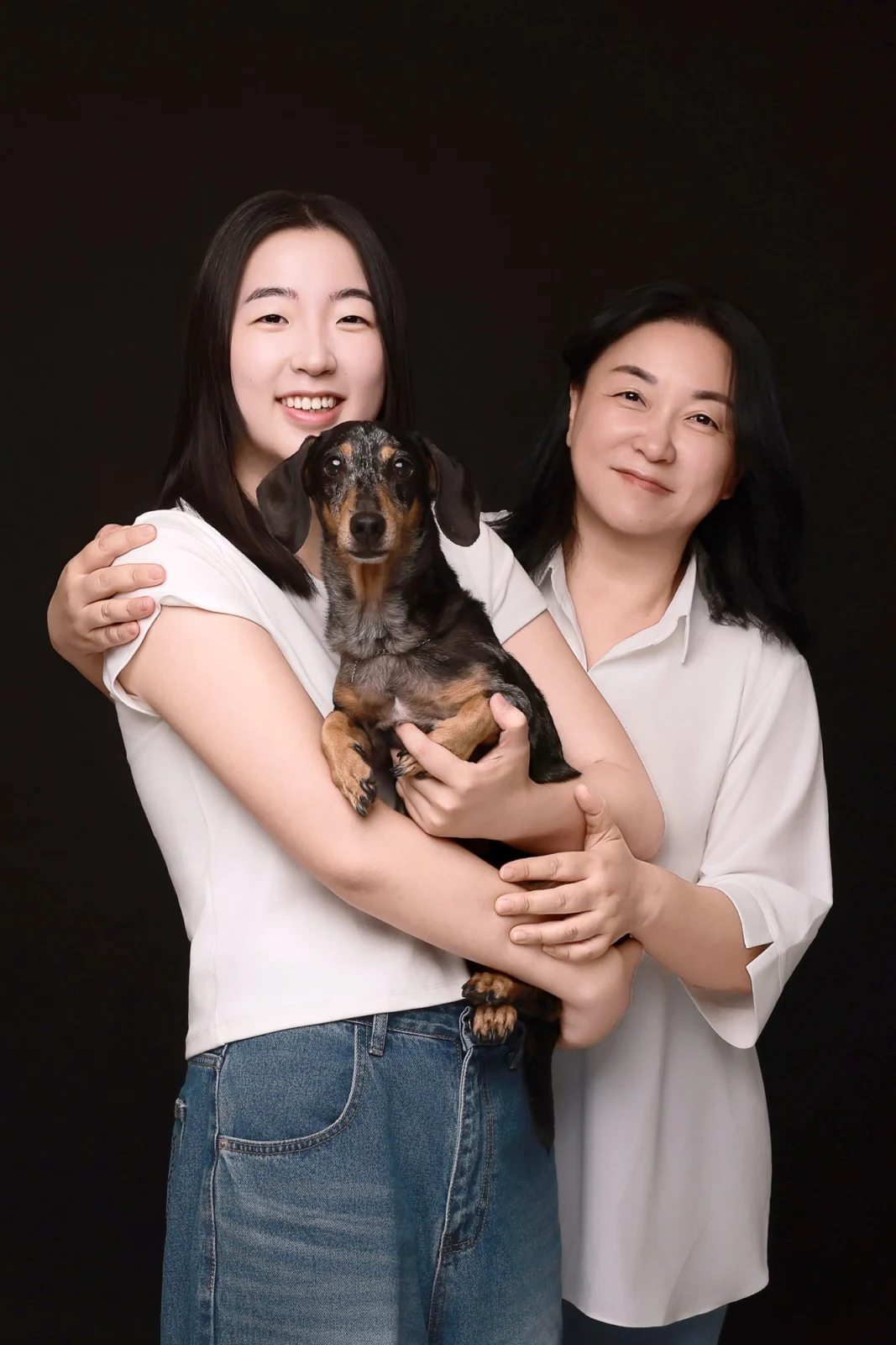 검정 배경에서 닥스훈트를 안은 사람을 옆 사람이 감싸 안은 두 사람의 개족사진