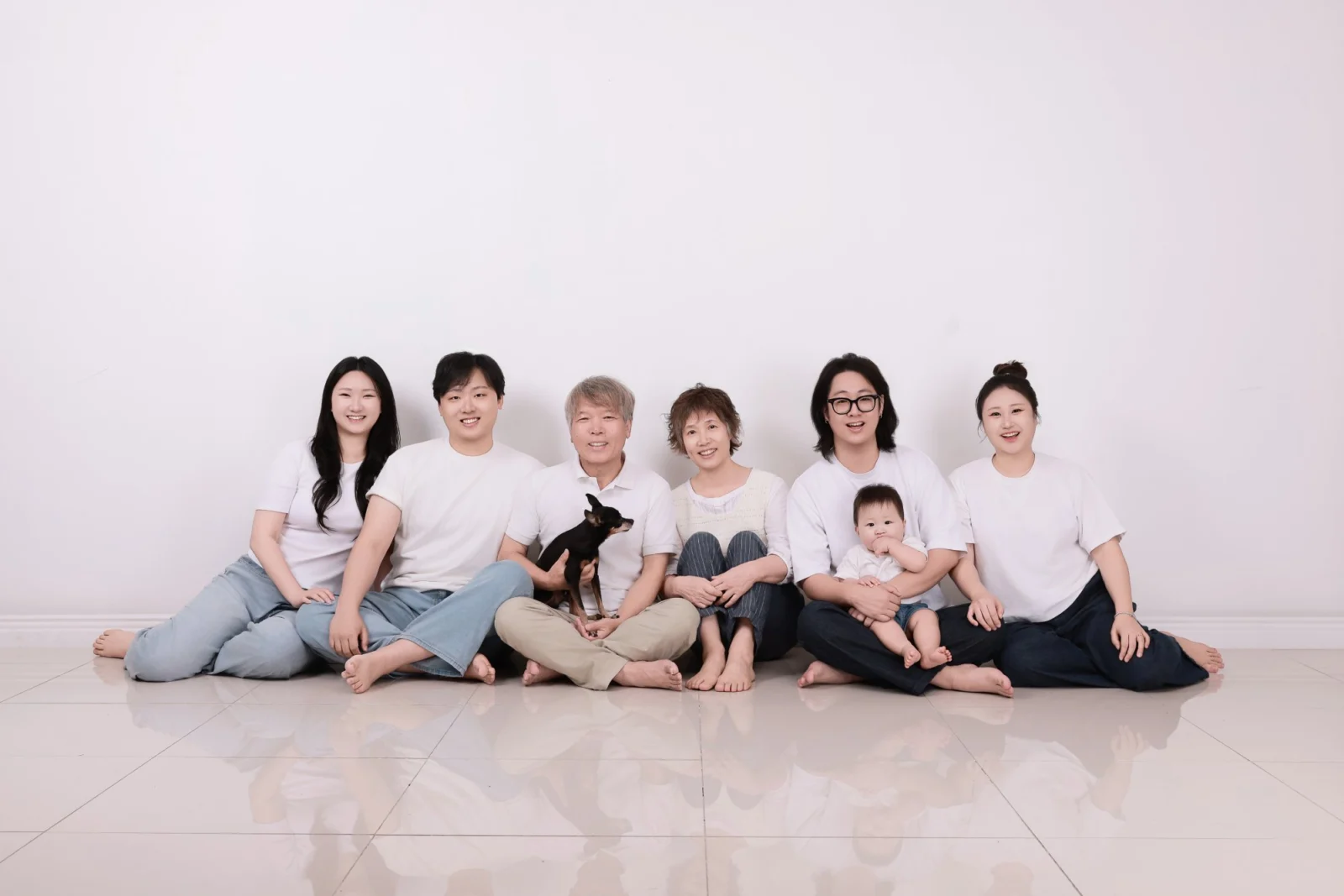 흰 바닥에 앉은 여섯 명이 아기와 검은 소형견을 안은 흰 티셔츠 개족사진