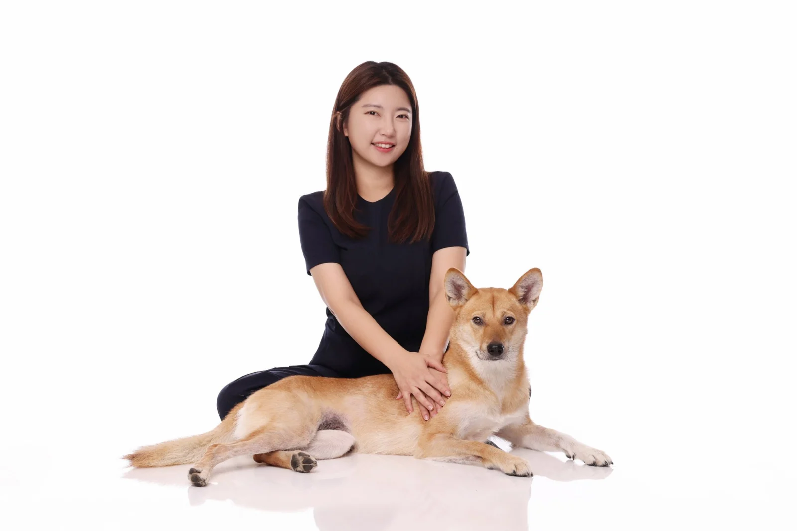 흰 바닥에 엎드린 갈색 중형견 뒤에 앉아 개를 쓰다듬는 한 사람의 둘이함개 사진