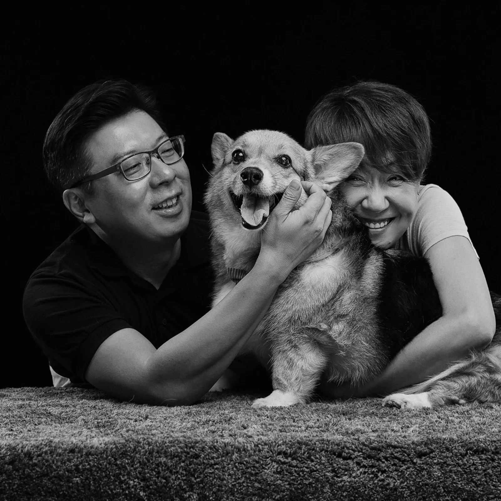 검정 배경 털 깔개 위 웰시코기를 끌어안고 웃는 두 사람의 흑백 가로 개족사진